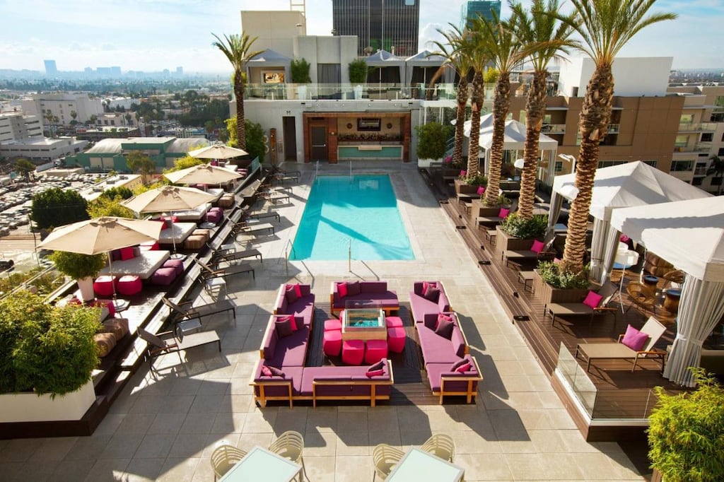 birds eye perspective of a cool Hollywood hotel rooftop pool and lounge area with pink sun furniture, white umbrellas and potted palm trees