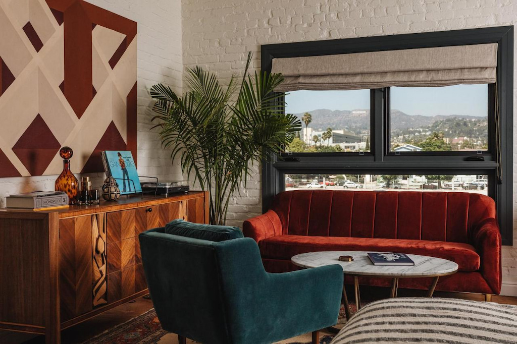 uber cool hotel in Los Angeles CA with red and blue living room furniture, wood side table, white brick walls, geometric art and window overlooking the city