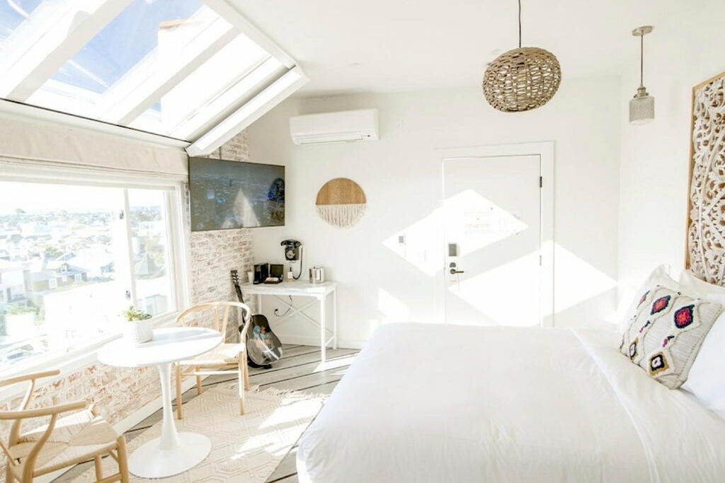 An all white room with a cozy bed, tables and chair and a flat screen TV and wooden decors near the window at a cool Venice Beach hotel