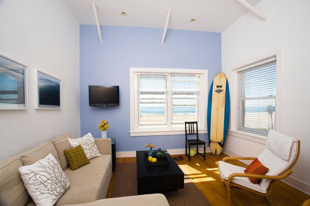 A small living room with a sofa, a center tale, an accent chair and a surfboard in the corner of the room near the window.