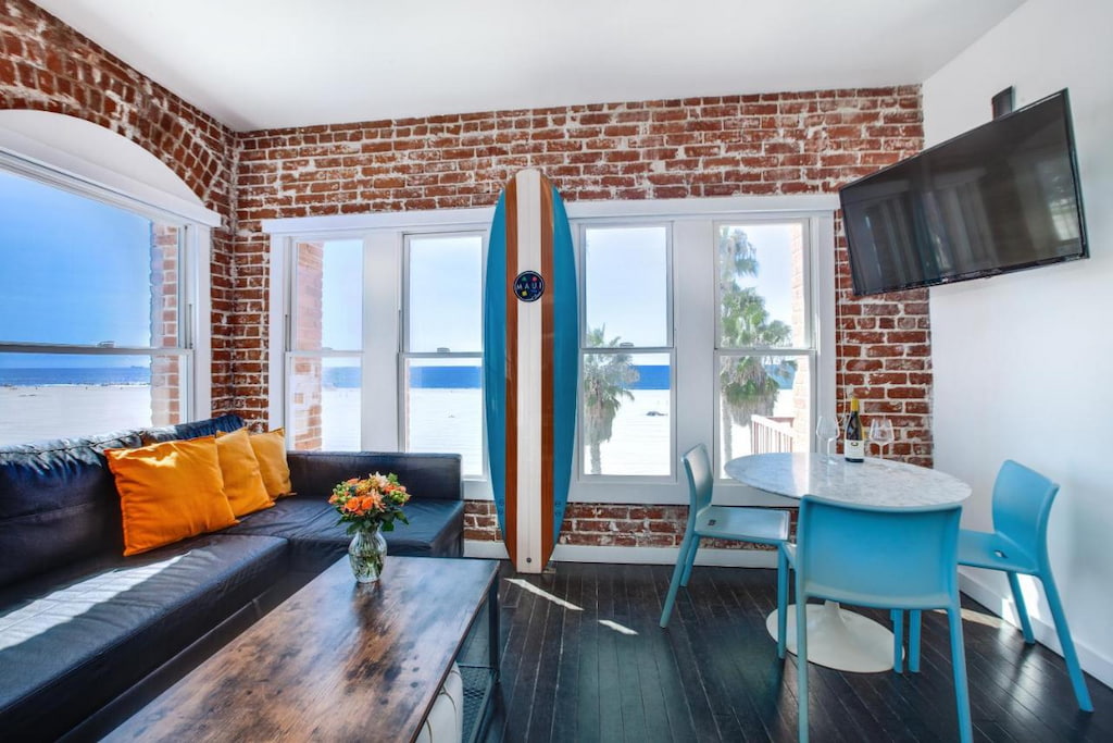 View of the living room area with a sofa with orange pillows, surfboard, rectangular coffee table and a blue dining set in a one of the coolest Venice Beach hotels.