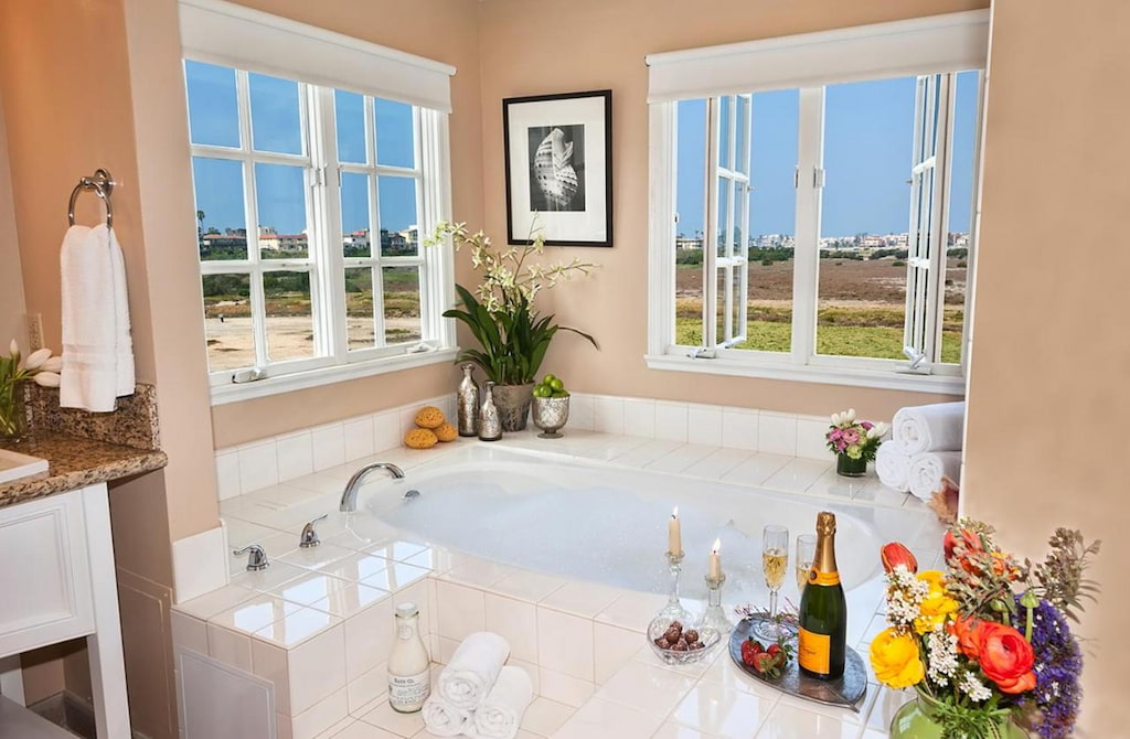 A romantic hotel in Venice Beach with a tub and a couple of windows with complimentary wine  bottles and flowers.