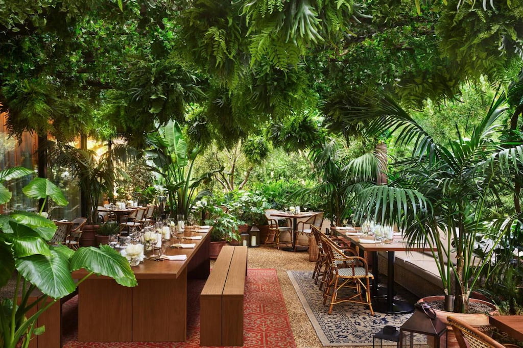An outdoor restaurant surrounded by greenery with rustic wooden tables and chairs at a cool West Hollywood hotel
