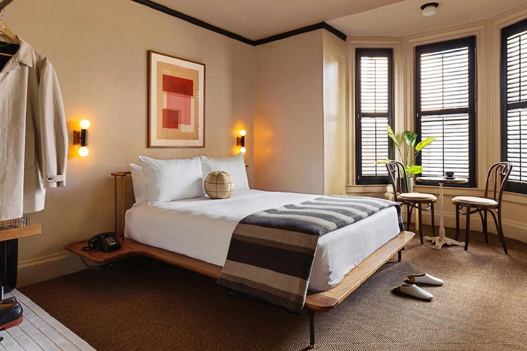 earthy toned boutique hotel in San Francisco with standard double bed and striped blanket, slippers and 3 black framed bay windows