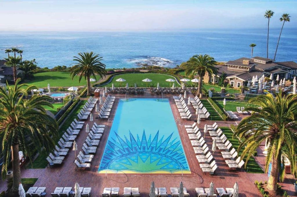 large outdoor pool area with half sun design, lined with white loungers, green space and Laguna Beach in the distance