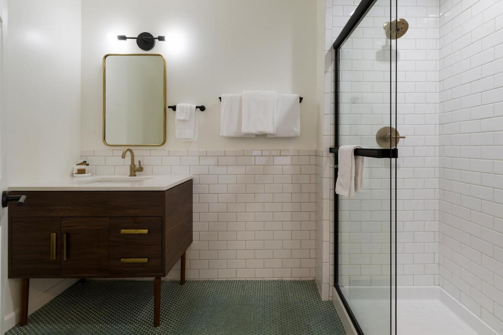 A cool hotel in Oakland bathroom with a shower with a glass divider and covered with white tiles, a sink with faucet and drawer, and a mirror