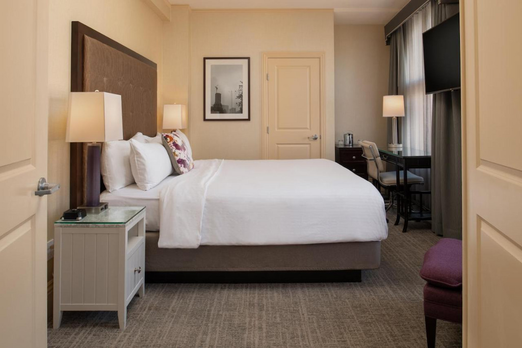 A carpeted hotel room with brown bed frame, white bed, bedside table with lamp, and desk with chair and lamp in one of the hotels UC Berkeley