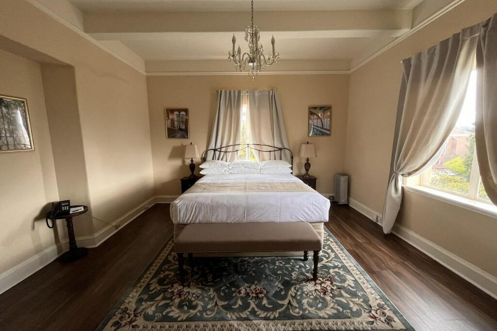 A suite with wooden floor, carpet, white bed, bedside tables with lamps, chandelier, and two curtained windows in one of the boutique hotels Berkeley CA
