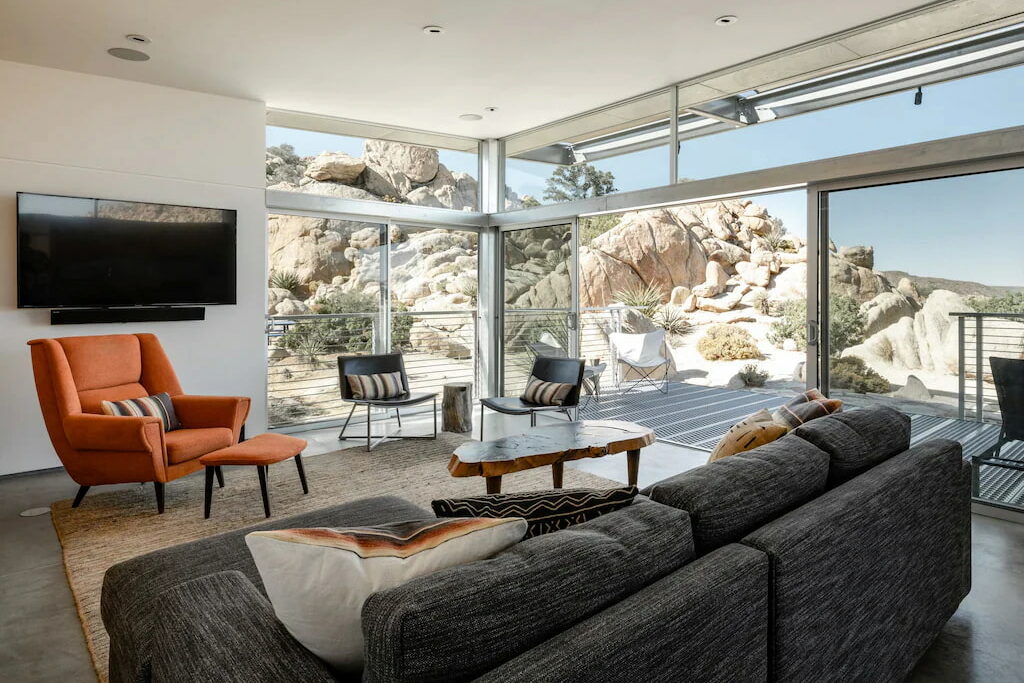 an orange accent chair in the corner of the room near the gray couch in front of ceiling-to-floor windows out to the desert