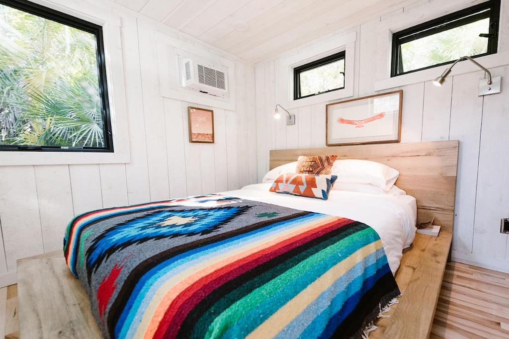 A bed with colorful striped blanket inside the caravan with white wood clad walls and black framed windows.