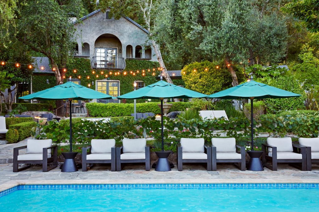 a charming boutique hotel in Sonoma Valley California in the background with a rectangular pool in the foreground with green umbrellas and white cushioned lounge chairs amongst greenery and twinkling lights.