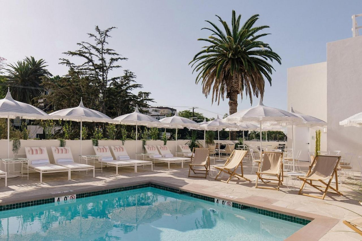 An outdoor swimming pool surrounded by a white building, trees, chairs, and umbrella shades