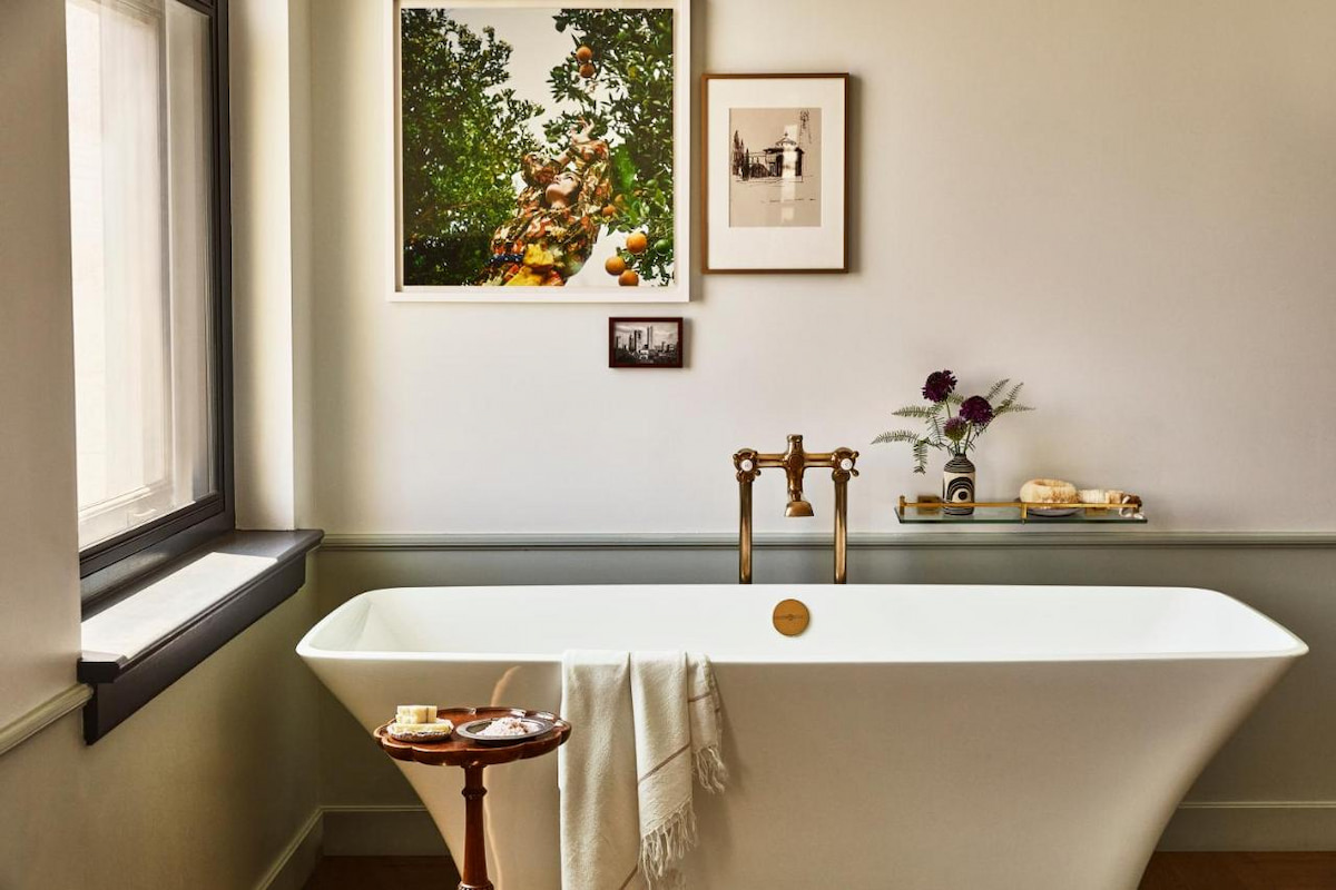 A bathroom in one of the coolest hotels in Southern California with a luxurious white bathtub, small wooden table, framed photographs, and window
