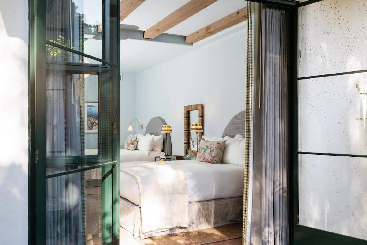 A view of a boutique hotel bedroom from the outside with two white beds, lamps, mirror, and floor-to-ceiling curtains