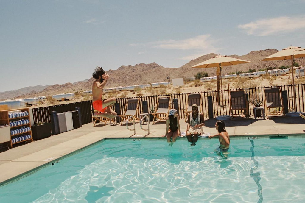 a man wearing bright red swim shorts jumps into an outdoor pool with desert surroundings
