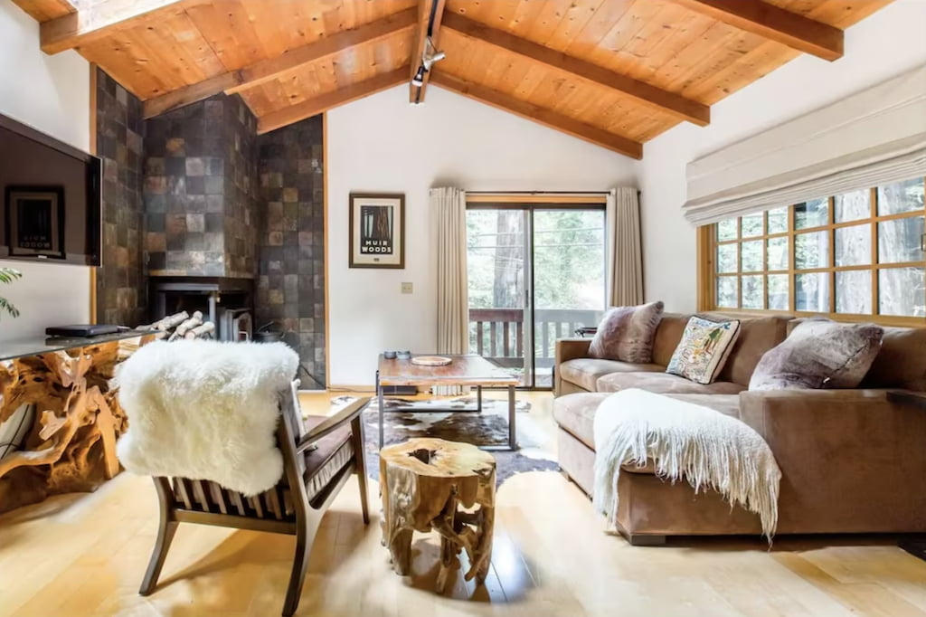 Luxurious cabin in Big Sur with brown sofa near the console table with wood bark legs, peaked wood ceiling and stone fireplace