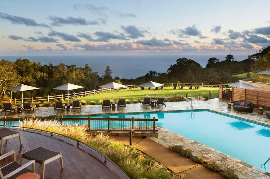 Panoramic of the ocean beyond a irregularly shaped outdoor pool with white umbrellas and greenery at dusk