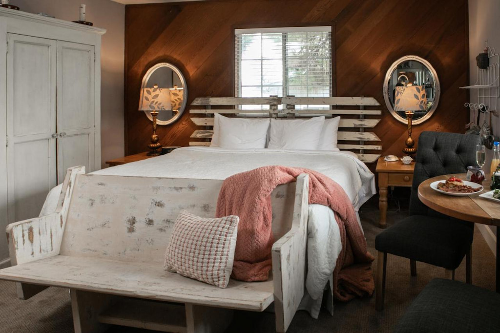 cozy wood cabin interior with white bed, rustic white bench with pink throw blanket and feature dark wood wall in a luxury hotel in Cambria California