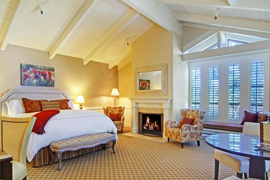 Carmel hotel room with queen-size bed, fireplace, tables, chairs, lamps, and white, wooden ceiling