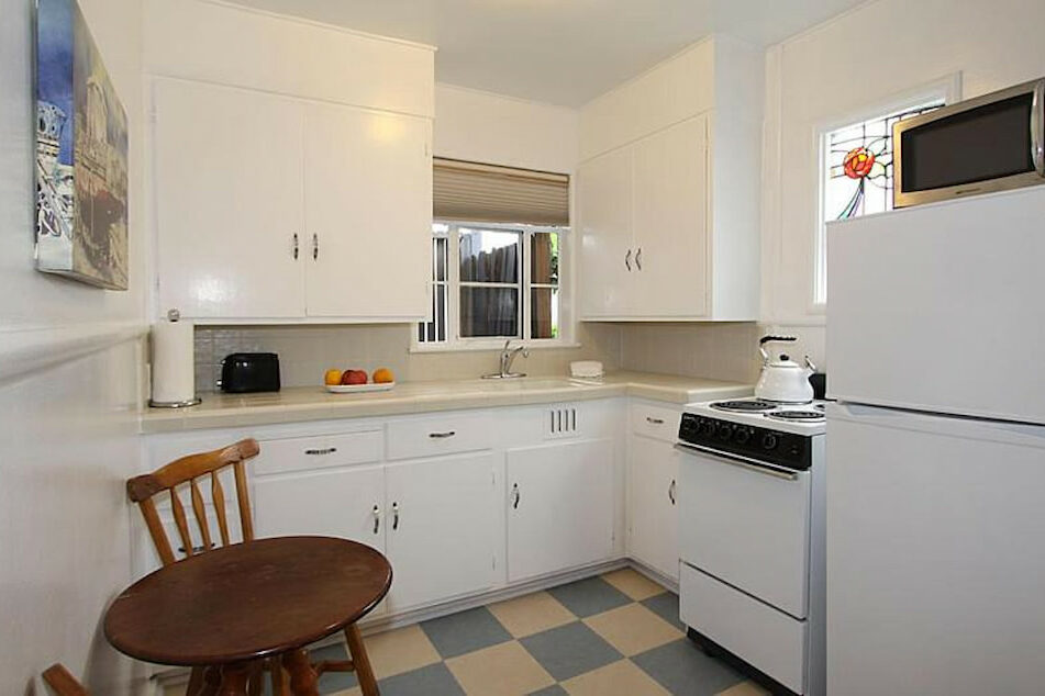 A white kitchenette with a counter, sink, cupboards, cooking range, refrigerator, microwave, and wooden table and chair