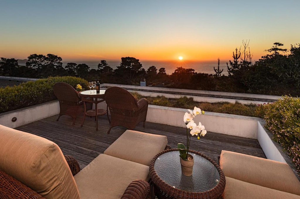 Outside sitting area with tables and chairs, surrounded by bushes and trees, and has a view of the sea at sunset