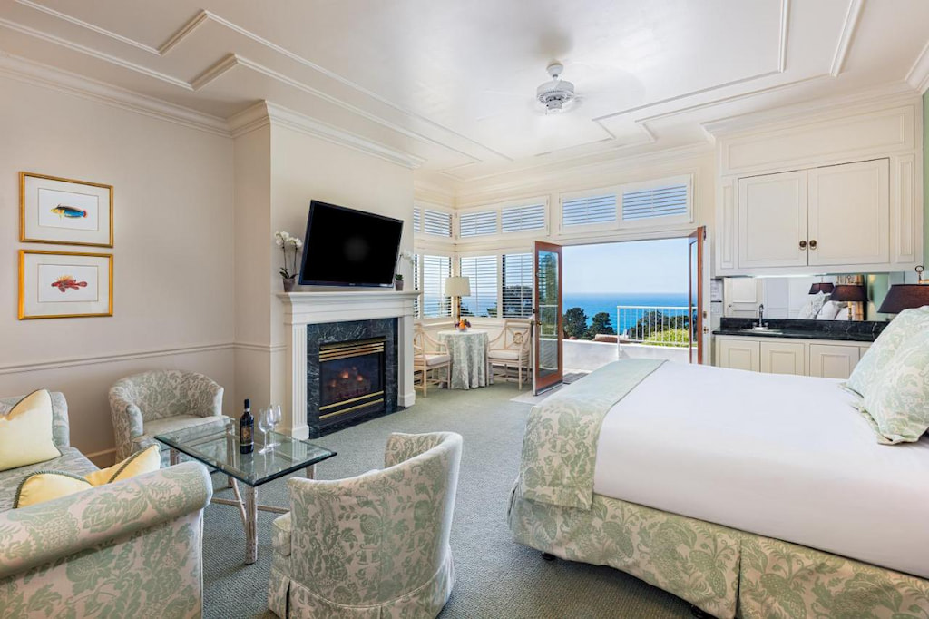 A carpeted hotel room with double bed, tables, chairs, flat-screen television over a fireplace, counter with cupboards, and with a view of the blue sea outside