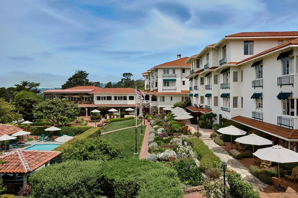 a three story white building with red roof surrounds a grassy courtyard with white umbellas and small pool at one of the best Carmel by the Sea boutique hotels.