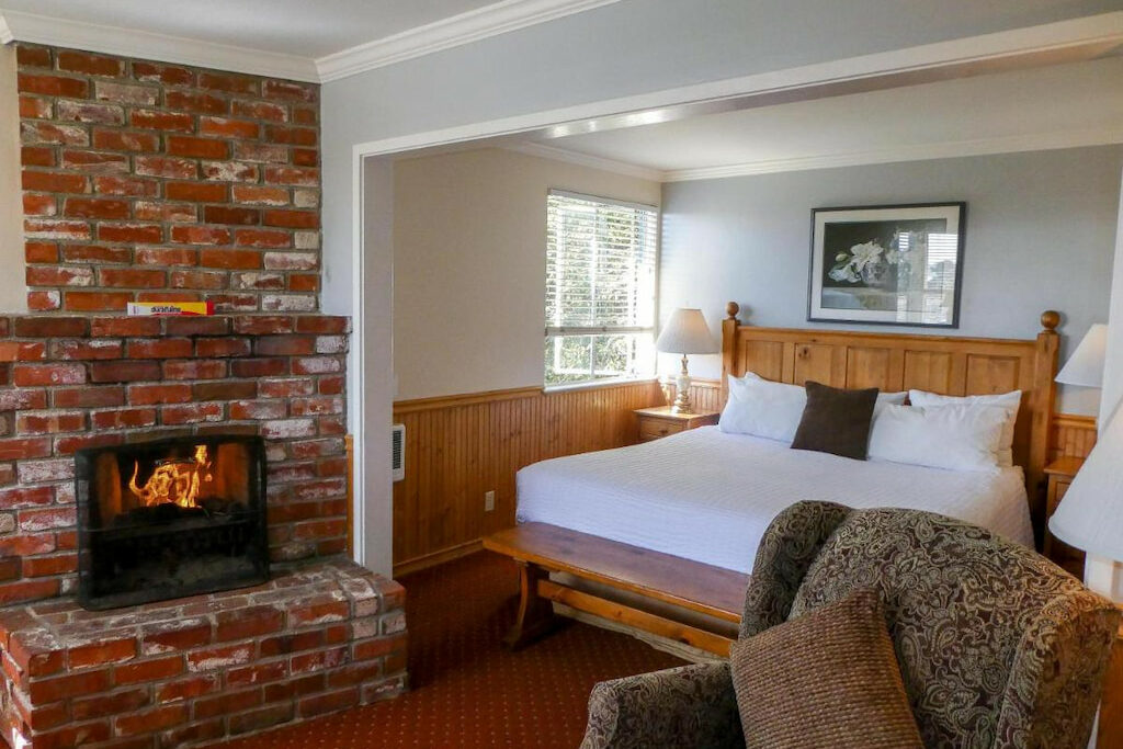 A room in a cottage in Carmel by the sea with white bed, bedside tables with lamps, upholstered chair, and brick fireplace