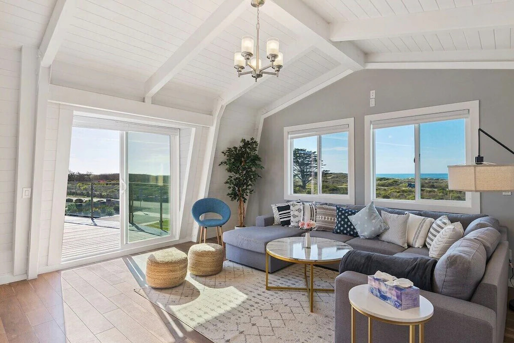 A Half Moon Bay California holiday home with a long gray couch, wooden ottoman and accent chairs and cute round tables near the glass doors.