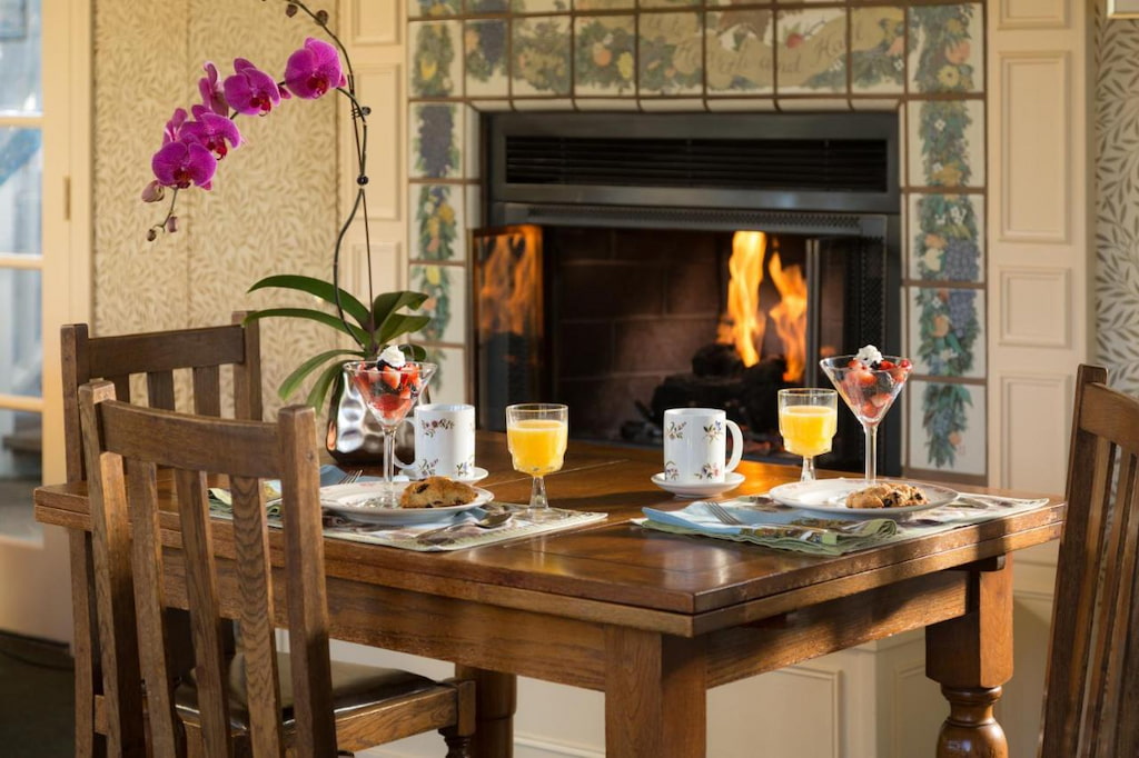 A fireplace beside a wooden dining set with a glass of juice and a yummy breakfast with an orchid flower on the center of the table.
