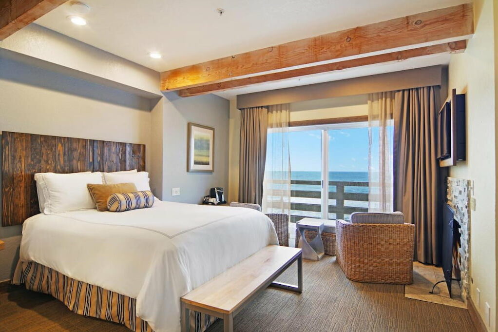 An Half Moon Bay boutique hotel room with wood design with a cozy bed with white sheets, a bench and a unique chair facing the sliding door with view of the beach.