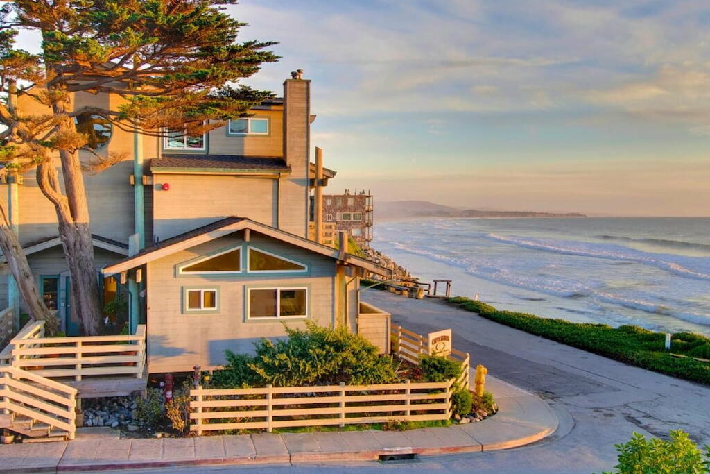 Exterior view of one of the luxury hotels in Half Moon Bay with a small fence and a tall tree near a small road and view of the beach.
