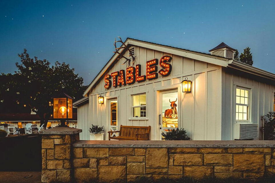 a boutique hotel in Paso Robles California at nighttime with white facade, stone fence and big red letters that say STABLES