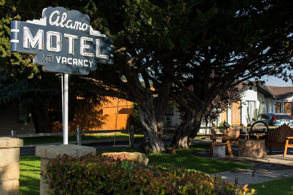large vintage roadside sign read Alamo Motel vacancy amongst large shrubbery, tree and green lawn with a unique hotel in Santa Ynez in the background