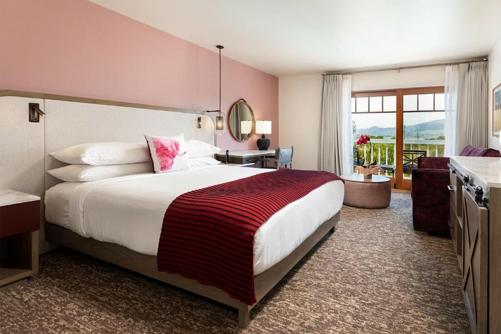 pink hotel room with large king sized bed with dark red throw blanket, a patio beyond large windows sits in the distance