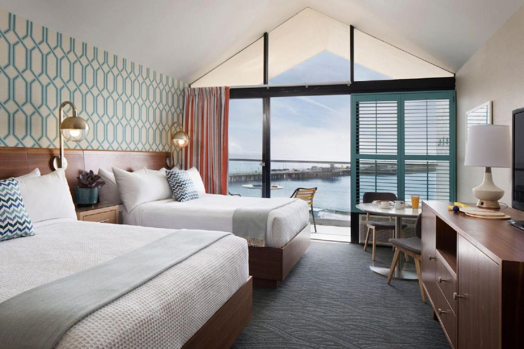 modern retro hotel room with peaked ceiling, wood console table and two beds with light linens with patio and ocean in the distance